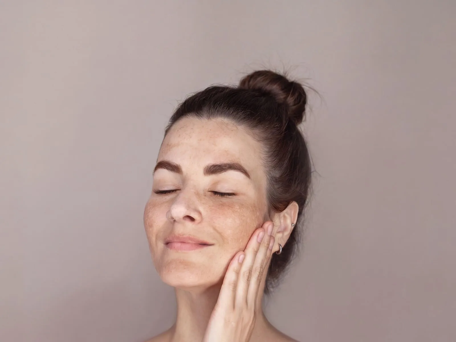 A close-up of a woman with her eyes closed and her fingertips resting against her cheek, freckling and soft brown melasma patches spread across both cheeks, the bridge of her nose and her upper lip