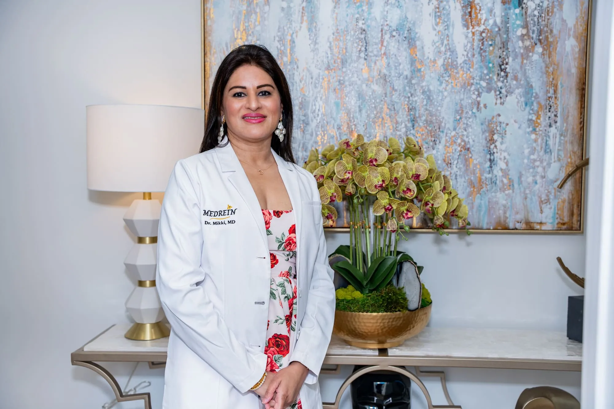 Dr. Haritha Mikkilineni, MD, in her Medrein lab coat beside the orchid display at the Medrein Health clinic in Southlake, TX