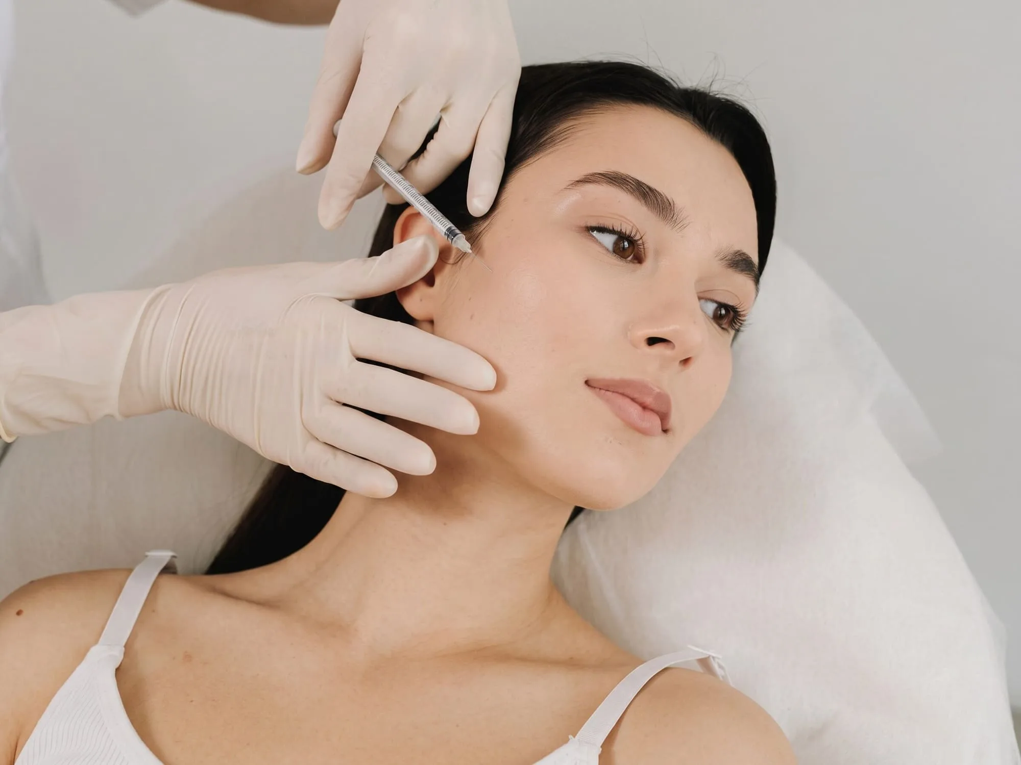 A gloved injector steadying a reclining patient's cheek while placing a fine-gauge syringe at her upper cheek in a bright treatment room