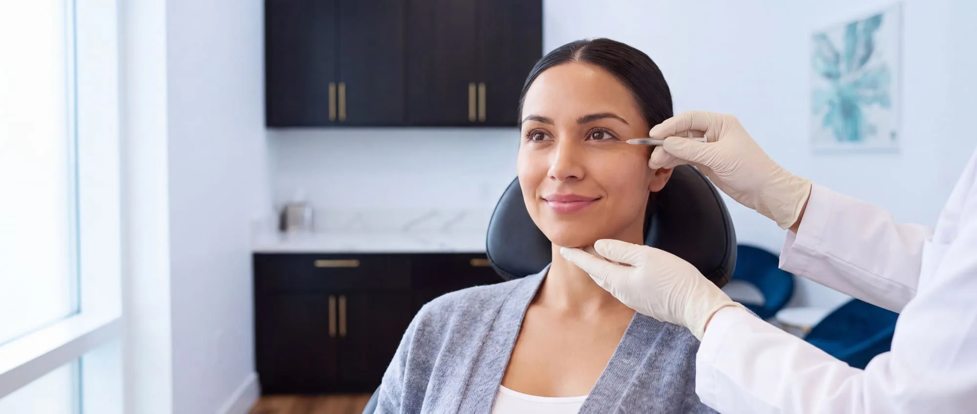 A seated patient smiling while a gloved clinician holds a fine-gauge syringe at her outer brow, the area neurotoxins treat