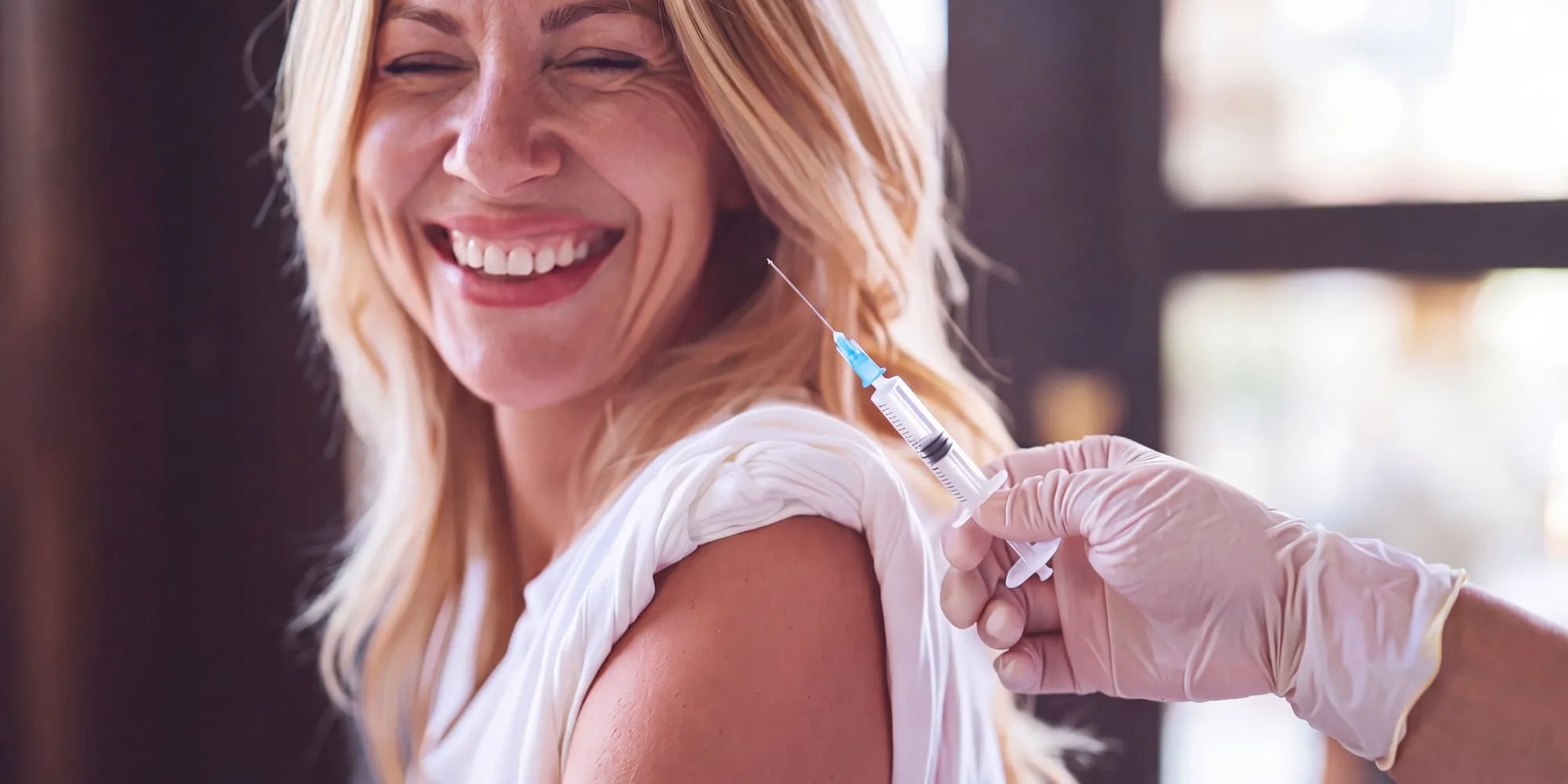 A woman laughing as a clinician in white gloves gives her a peptide injection in the outer upper arm, in a sunlit treatment room