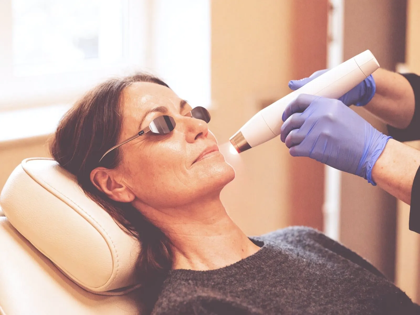 A handheld laser applicator held just above a reclining patient's jaw, a small pool of low-level light falling on the skin beneath its tip, with protective eye shields in place
