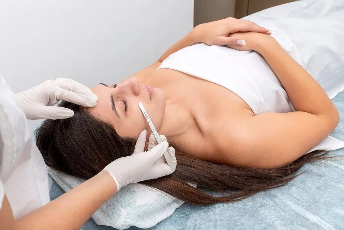 Gloved hands drawing a sterile dermaplaning blade at an angle across the cheek of a reclining patient