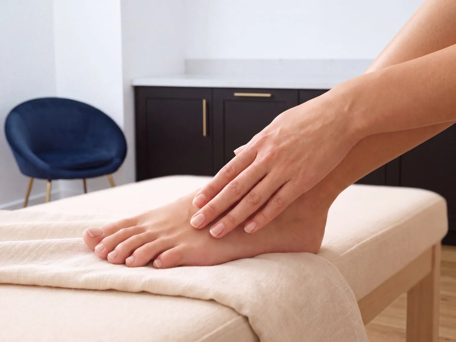 A well-groomed hand resting on a bare foot in a softly lit treatment room with cool white walls, dark cabinetry and a navy velvet chair, with clear, healthy fingernails and toenails both in view. Illustrative photograph, not a patient, and not a before or after.
