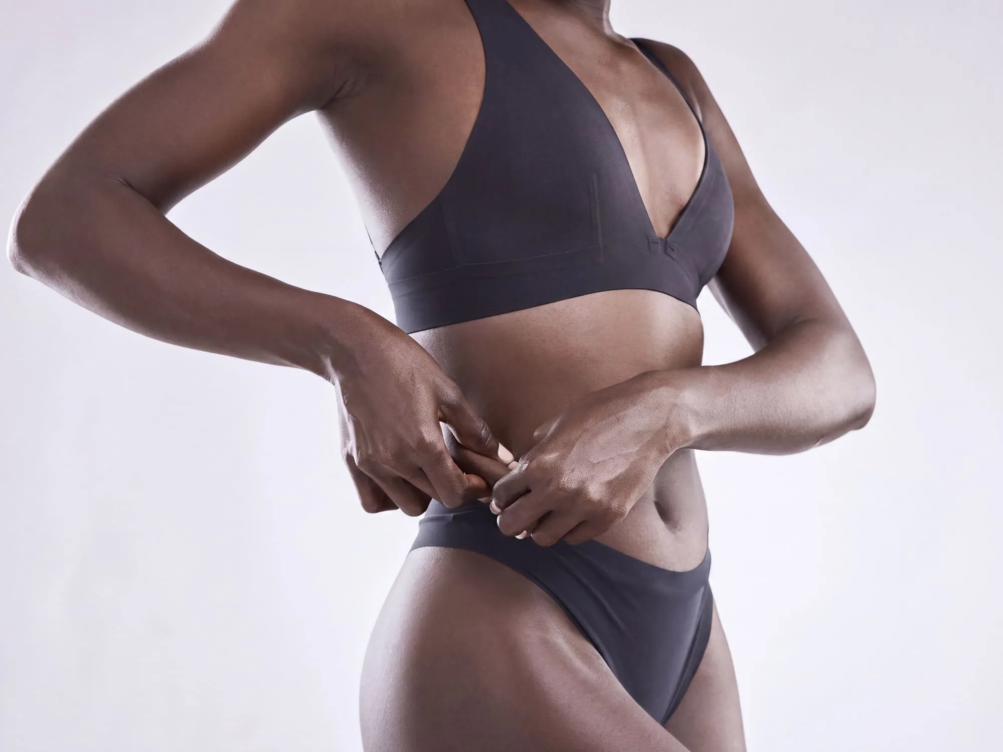 A woman in a charcoal bra and briefs against a pale grey studio background, both hands resting at her waist