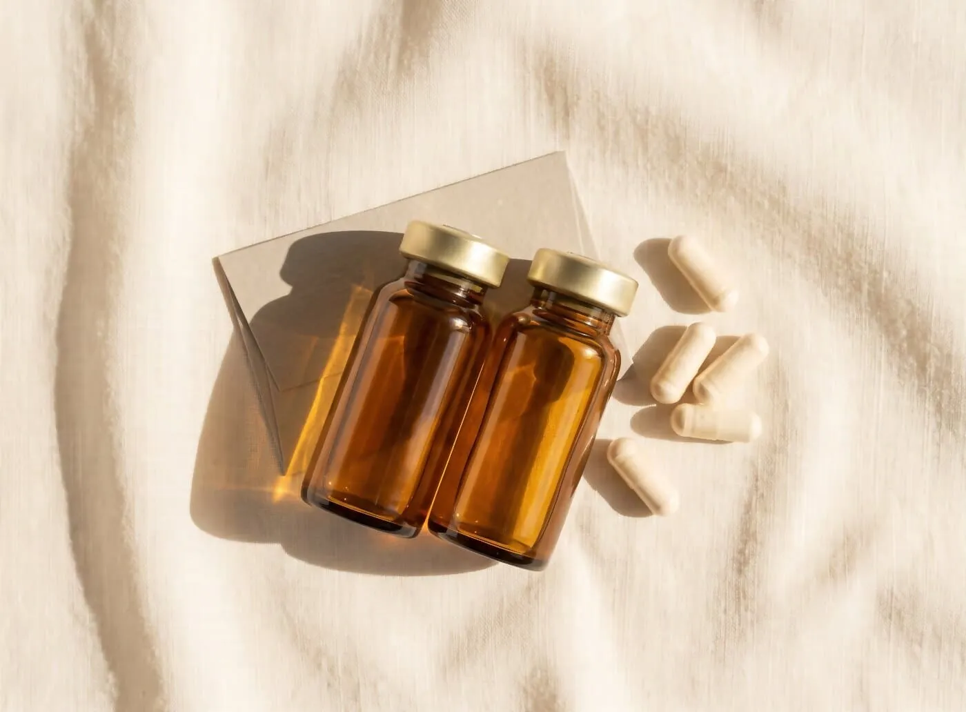 Two amber glass medication vials with crimped gold caps beside five pale capsules on warm cream linen in raking daylight