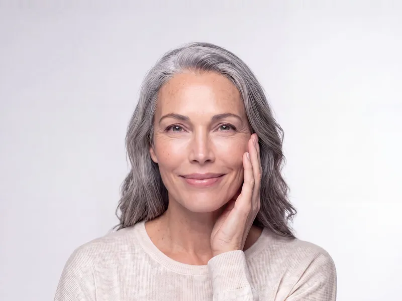 A woman with long silver hair resting her fingertips against her cheek and smiling.