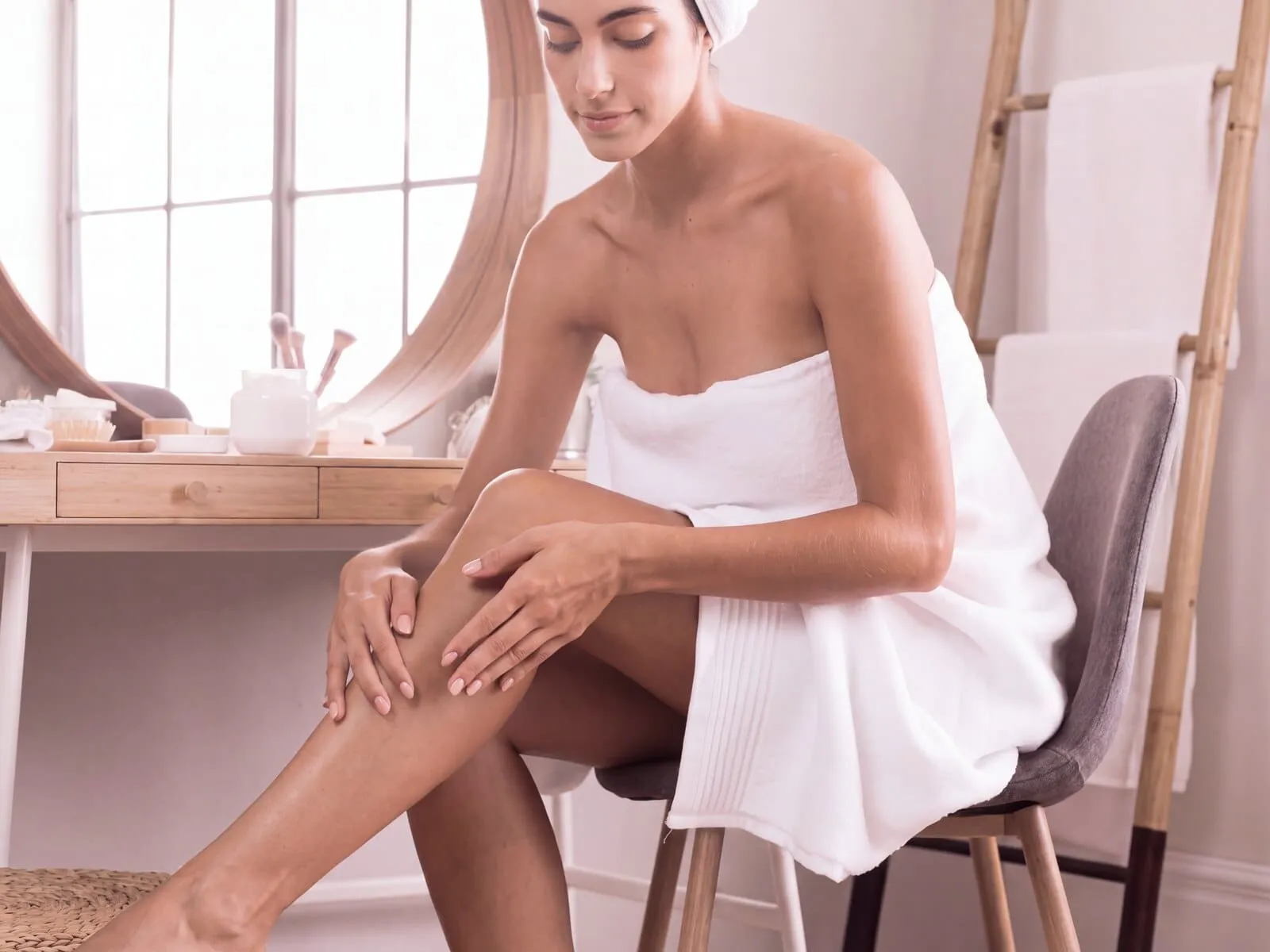 Woman wrapped in a towel after a shower, seated in a bright bathroom and running her hands over her smooth lower leg, one of the areas laser hair removal treats