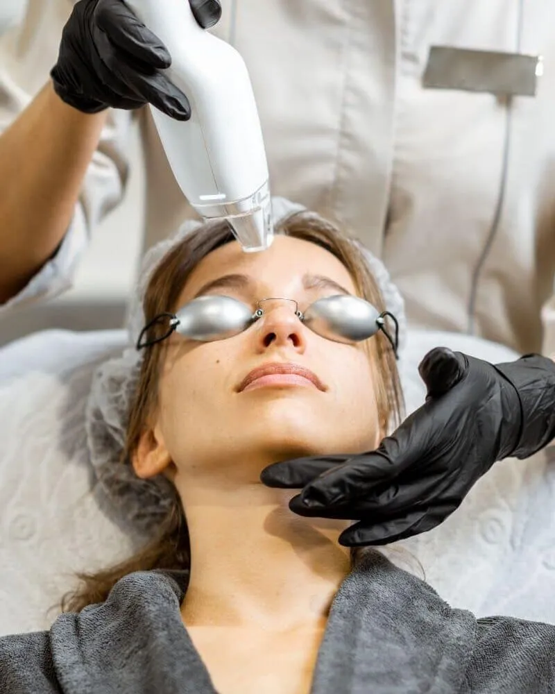 A clinician passing a pico laser handpiece over a reclining patient's face, protective eyewear in place, during a pigment treatment
